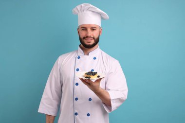 Üniformalı mutlu pastacı açık mavi arka planda yaban mersini tartı tutuyor.