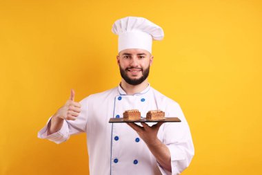 Üniformalı mutlu pastacı elinde çikolatalı tatlılar ve turuncu arka planda baş parmağını kaldırıyor.