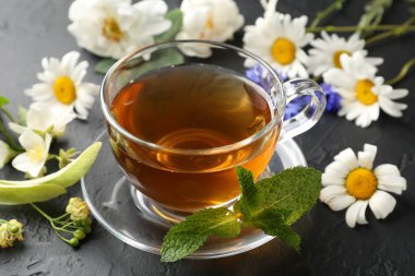 Cam fincanda aromatik bitki çayı, farklı çiçekler ve siyah masada nane şekeri, yakın plan.