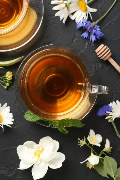 Cam fincanlarda aromatik bitki çayı, farklı çiçekler ve siyah masada nane şekeri.