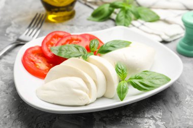 Lezzetli mozzarella peyniri, domates, fesleğen ve baharatlar gri masada, yakın plan.