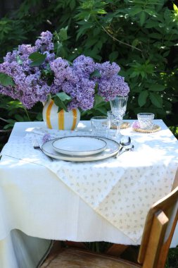 Bahçedeki masada güzel leylak çiçekleri, tabaklar, çatal bıçaklar, bardaklar ve mumlar