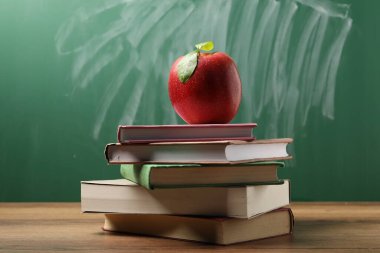 Tahtanın yanındaki ahşap masada farklı kitaplar ve kırmızı elma, yakın plan.