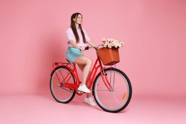 Gülümseyen kadın pembe arka planda çiçeklerle bisiklet üstünde poz veriyor.