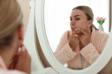 Evde aynaya bakan sivilceli bir kadın.