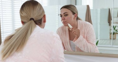 Kadın evin içinde aynanın yanında yüzüne krem sürüyor. Sivilce tedavisi