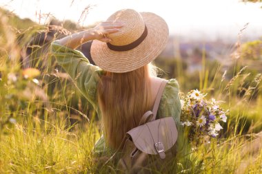 Güzel kır çiçekleriyle, şapkasıyla sırt çantasıyla güneşli bir günde çayırda gezen bir kadın.