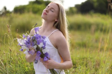Bir buket kır çiçeğiyle güzel bir kadın çayırda güneşli bir günün tadını çıkarıyor.