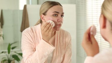 Sivilceli bir kadın aynanın içinde yüzünü temizliyor.