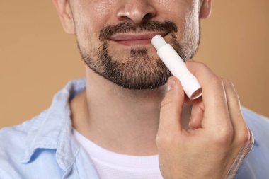 Açık kahverengi arka planda dudaklarına dudak kremi süren adam, yakın plan.