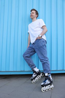 Açık mavi duvarın yanında patenleriyle gezen mutlu genç bir adam.
