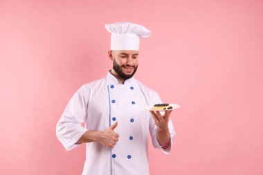 Üniformalı mutlu pastacı elinde yaban mersinli turta ve pembe arka planda baş parmağını gösteriyor.