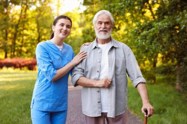 Gülen hemşire dışarıda bastonuyla yaşlı bir adamı destekliyor.