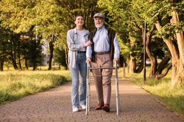 Gülümseyen bakıcı dışarıda yürüyen çerçeveyi kullanan yaşlı bir adamı destekliyor.