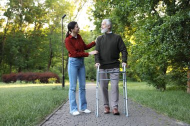 Gülümseyen bakıcı dışarıda yürüyen çerçeveyi kullanan yaşlı bir adamı destekliyor.