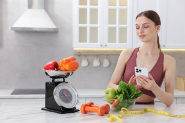 Beyaz mermer masanın yanında akıllı telefon kullanan bir kadın. Mutfak terazisi, sağlıklı yiyecekler, dambıllar ve kapalı alanda mezura kullanıyor.