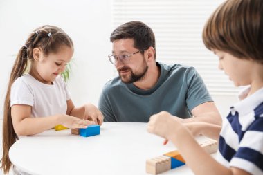 Babam ve çocukları içeride beyaz masada tahta figürlerle oynuyorlar.