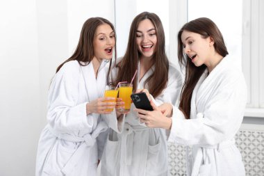 Bornozlu ve portakal suyu bardaklı mutlu kadınlar spa salonundaki akıllı telefonlara bakarlar.