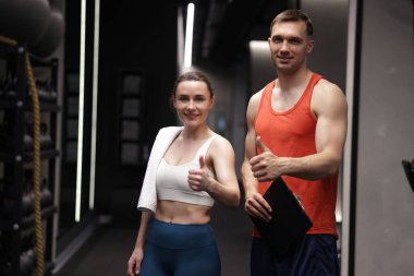 Profesyonel kişisel antrenörü olan genç bir kadın jimnastik salonunda baş parmağını kaldırıyor.