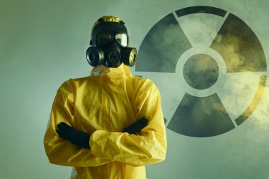 Tehlikeli madde giysisi giymiş ve gaz maskesi takmış bir adam. Arkasındaki renk arkaplanında radyoaktif işaret