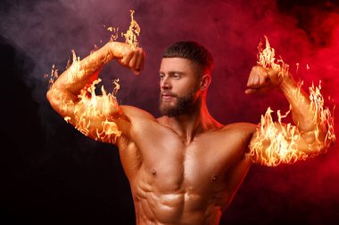 Vücut geliştirici karanlık arka planda pazılarını duman içinde gösteriyor. Alevli kollu kaslı adam