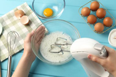 Kadın, açık mavi ahşap masada el çırpıcı ile yumurta beyazı çırpıyor.