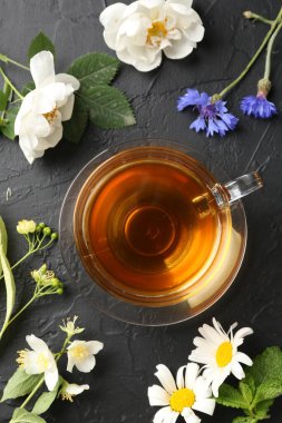 Cam fincanda aromatik bitki çayı, farklı çiçekler ve siyah masada nane şekeri.