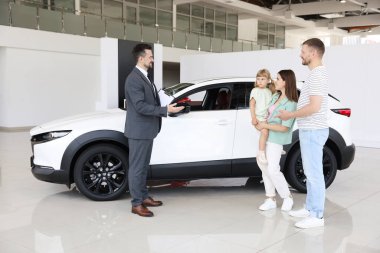 Gülümseyen satıcı elinde dosyayla salonda aileye yeni bir araba gösteriyor.