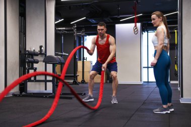 Profesyonel özel antrenör, genç bayana spor salonunda savaş halatlarıyla nasıl egzersiz yapılacağını gösteriyor.