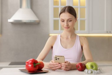 Beyaz masada akıllı telefon kullanan bir kadın. Mutfak terazisi, sağlıklı yiyecekler ve kapalı alanda mezura kullanıyor.