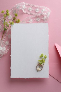 Düğün davetiyesi, zarf, altın yüzükler, dantel kurdele ve pembe arka planda çiçekli dallar.