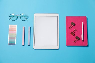 Tablet, farklı kırtasiye malzemeleri ve açık mavi arka planda gözlük, düz uzanma