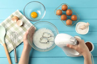 Bir kadın açık mavi ahşap masada el çırpıcı ile yumurta beyazı çırpıyor.