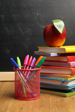 Tahtanın yanındaki ahşap masada farklı kırtasiye malzemeleri ve kırmızı elma.