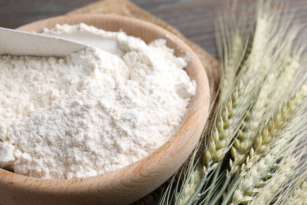 Flour in bowl and green wheat spikes on table, closeup