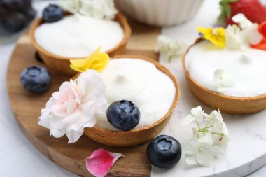 Lezzetli tatlı. Kremalı tartlar, böğürtlenler ve çiçekler beyaz döşemeli masada, yakın plan.