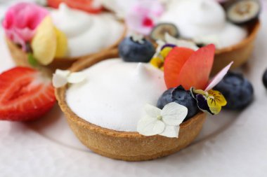 Lezzetli tatlı. Kremalı tartlar, böğürtlenler ve çiçekler beyaz döşemeli masada, yakın plan.