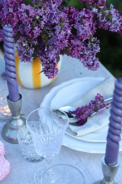 Güzel leylak çiçekleri, tabaklar, çatal bıçak takımı, cam ve mumlar bahçede, yukarıdaki manzaranın üzerinde.