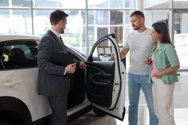 Elinde dosya olan satıcı salonda mutlu bir aileye yeni bir araba gösteriyor.