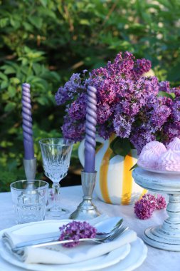Güzel leylak çiçekleri, zephyrler, tabaklar, çatal bıçaklar, bardaklar ve mumlar bahçede, yakın plan