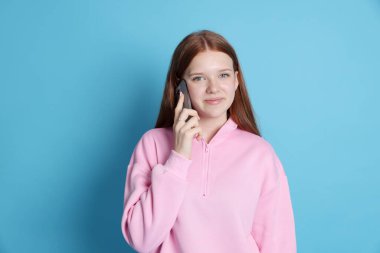 Güzel genç kız açık mavi arka planda akıllı telefondan konuşuyor.