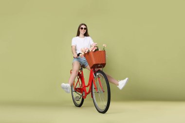 Güneş gözlüklü gülümseyen kadın yeşil arka planda bir sepet çiçekle bisiklet sürüyor.