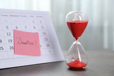 Kelime dağılımı, takvim ve kum saati içeren yapışkan bir not. İçerideki ahşap masada akan kum, yakın plan.