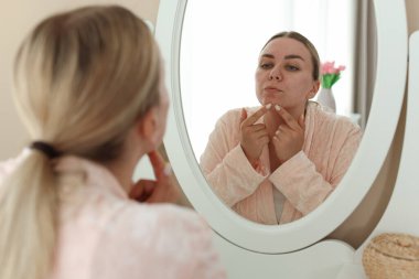 Evde aynaya bakan sivilceli bir kadın.