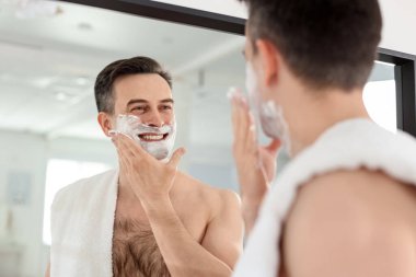 Gülümseyen adam evde aynanın yanında tıraş köpüğü sürüyor. Düşük açı.