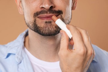 Açık kahverengi arka planda dudaklarına dudak kremi süren adam, yakın plan.