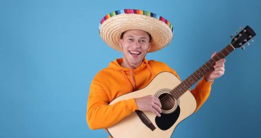 Şapkalı mutlu adam açık mavi arka planda gitar çalıyor.