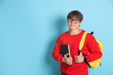 Açık mavi arka planda kitap ve sırt çantalı, gözlüklü, gülümseyen genç çocuk. Metin için boşluk