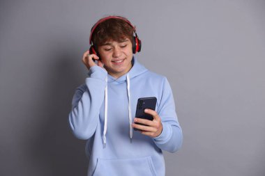 Gri arka planda akıllı telefon kullanan, kulaklıklı, gülümseyen genç çocuk.
