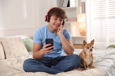 Evde akıllı telefon ve köpek kullanarak kulaklık takan gülümseyen genç bir çocuk.